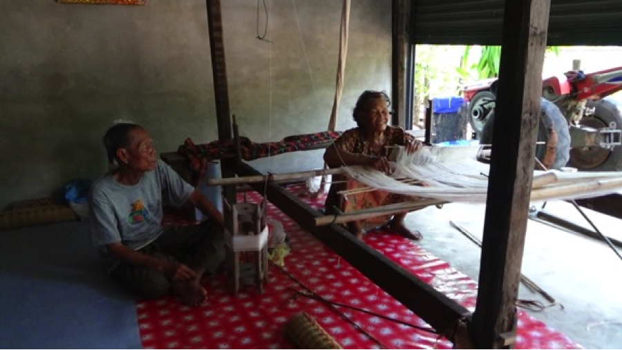As her husband looks on, Ms. Khamlamun Srikham passes the time weaving, no longer able to engage in farming.